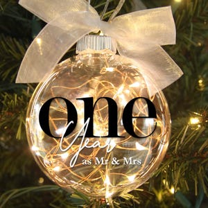 May include: A clear glass Christmas ornament with the words "one year as Mr & Mrs" in black and white script. The ornament is filled with small, warm white lights and is tied with a sheer white ribbon.