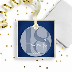 May include: A square navy blue ornament featuring a light blue circle with the number 18, the name Hannah in white script, and the year 2026. The ornament hangs from a cream-colored ribbon and is presented in a white gift box with gold confetti.