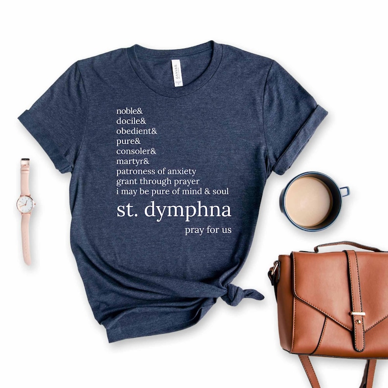 May include: A navy blue t-shirt with white text listing virtues and the words "st. dymphna pray for us." The shirt is styled with a knot at the bottom. A watch, coffee cup, and brown handbag are also in the image.