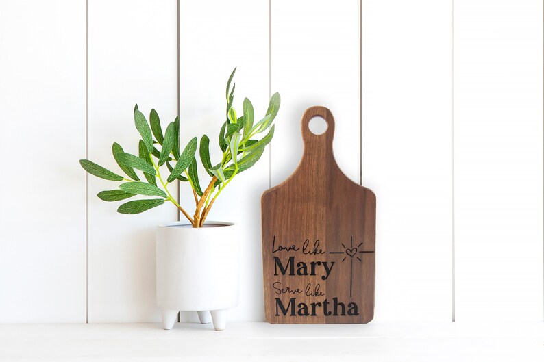 May include: A wooden cutting board with a handle, engraved with the text "Love like Mary, Serve like Martha" and a small heart with a cross.