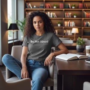 以下が含まれることがあります： 「Be Not Afraid」の白い文字がプリントされた濃いグレーのTシャツ。椅子に座っている人が着ています。その人はブルージーンズを履いています。本とコーヒーカップがテーブルの上にあります。