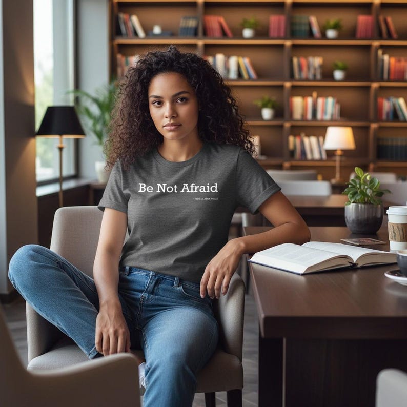 以下が含まれることがあります： 「Be Not Afraid」の白い文字がプリントされた濃いグレーのTシャツ。椅子に座っている人が着ています。その人はブルージーンズを履いています。本とコーヒーカップがテーブルの上にあります。