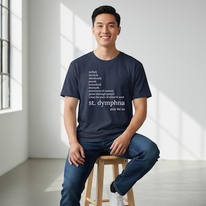 May include: A navy blue t-shirt with white text. The text includes the words "noble," "docile," and "St. Dymphna." The shirt is worn by a person sitting on a wooden stool. The person is wearing blue jeans.