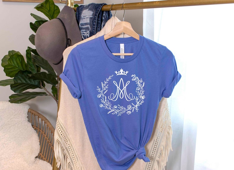 May include: A periwinkle blue t-shirt with a white floral wreath design, a crown, and the letter "M". The shirt is hanging on a wooden hanger. Rolled sleeves and a tied knot at the bottom.