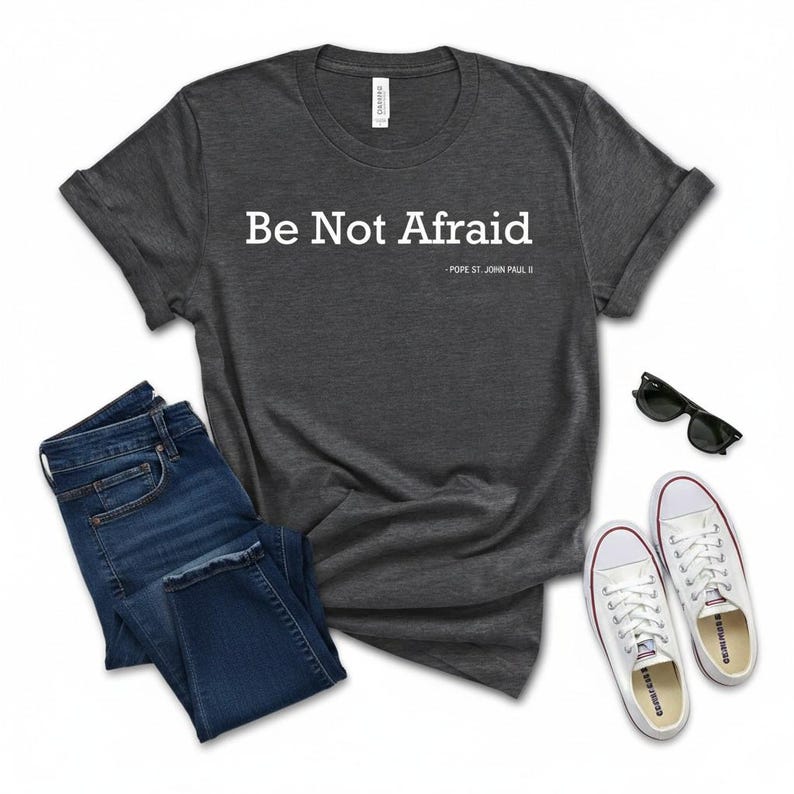 以下が含まれることがあります： 「Be Not Afraid」と「- Pope St. John Paul II」の文字が入った濃いグレーのTシャツ。青いジーンズ、白いスニーカー、黒いサングラスも画像にあります。シャツは折りたたまれています。