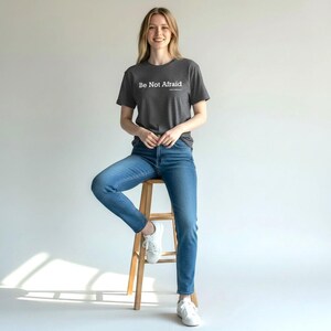 以下が含まれることがあります： 「Be Not Afraid」の白文字がプリントされた濃いグレーのTシャツ。青いジーンズと白いスニーカーを合わせています。木製のスツールに座っています。