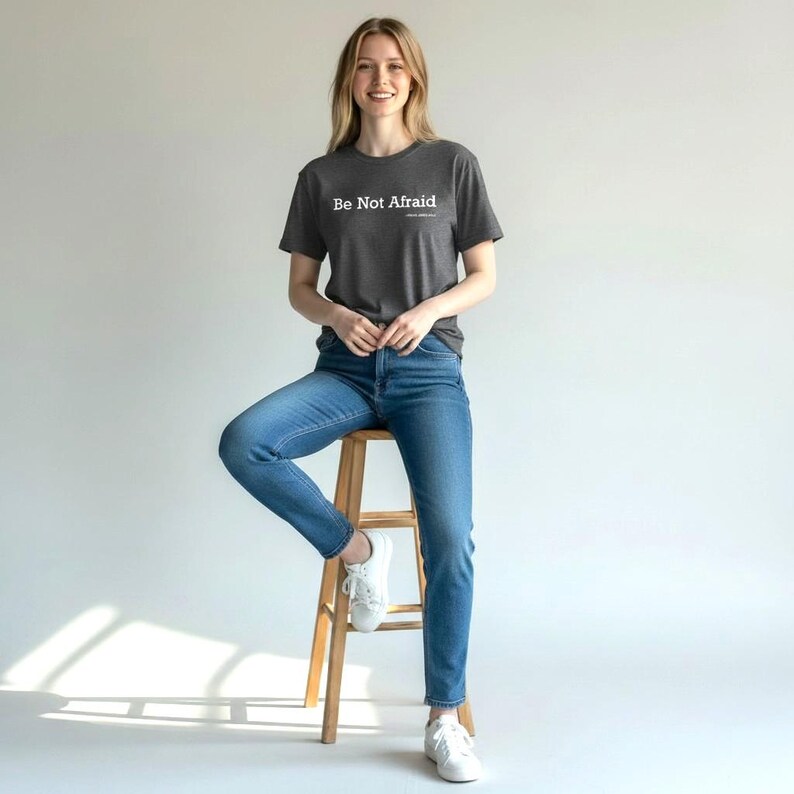 以下が含まれることがあります： 「Be Not Afraid」の白文字がプリントされた濃いグレーのTシャツ。青いジーンズと白いスニーカーを合わせています。木製のスツールに座っています。