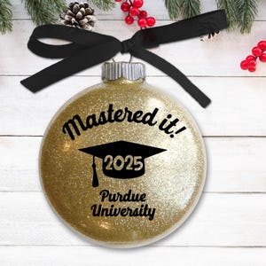 Peut inclure: Boule de Noël dorée à paillettes avec un bonnet de graduation noir et le texte "Mastered it! 2025 Purdue University".