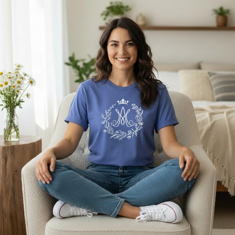 May include: A blue t-shirt featuring a white floral wreath design with a crown and the letter "A". The shirt is made of a soft material and is being worn.