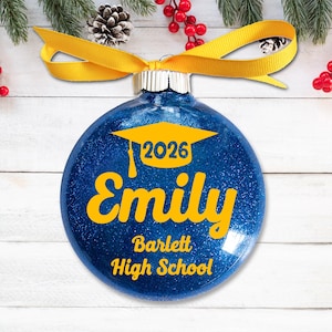 May include: A blue glitter Christmas ornament with a yellow ribbon. The ornament has the name "Emily", the year "2026", and "Barlett High School" in yellow text, along with a graduation cap graphic.