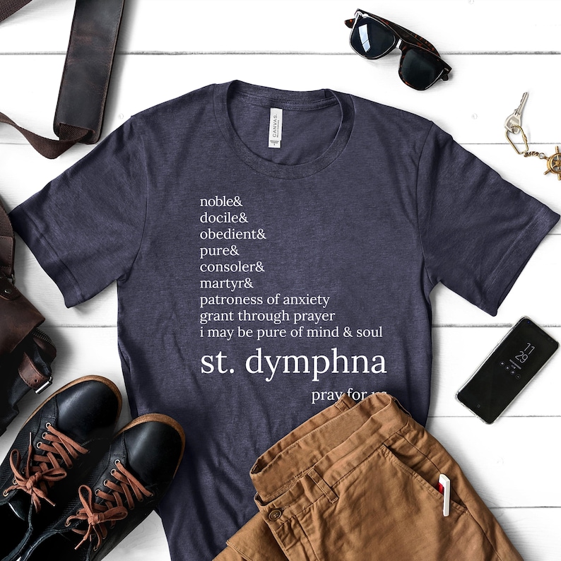 May include: A navy blue t-shirt with white text listing virtues and the words "st. dymphna pray for." The shirt is displayed with brown trousers, black shoes, sunglasses, and a phone on a white wooden surface.