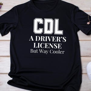May include: Black t-shirt with white text that reads "CDL A DRIVER'S LICENSE But Way Cooler". The letters "CDL" are in a bold, outlined font. The shirt is made of a soft, dark fabric.