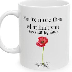 Könnte beinhalten: Ein weißer Keramikbecher mit schwarzem Text, der lautet: You're more than what hurt you, There's still joy within. Unter dem Text befindet sich eine Aquarellillustration einer einzelnen roten Mohnblume, die aus einem Riss im Boden wächst.
