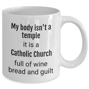 May include: White ceramic coffee mug with black text that reads "My body isn't a temple it is a Catholic Church full of wine bread and guilt"