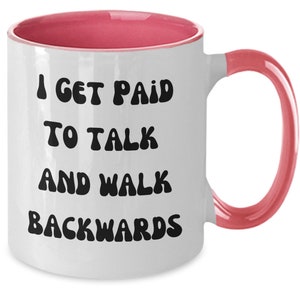 Peut inclure: Une tasse à café en céramique blanche avec un bord et une poignée roses. La tasse porte une inscription noire qui dit "Je suis payé pour parler et marcher à reculons".
