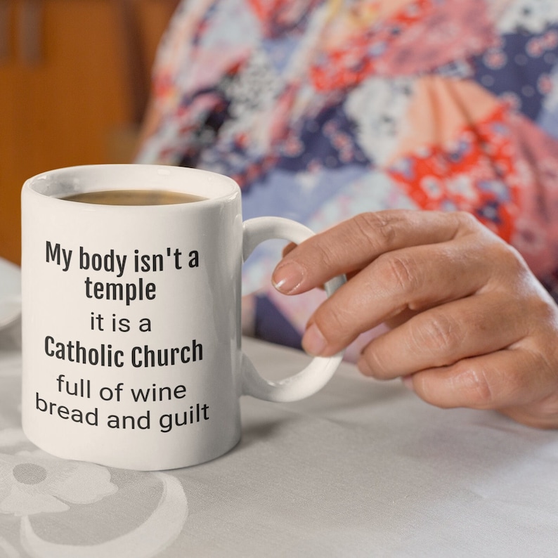 May include: White ceramic mug with black text that reads "My body isn't a temple it is a Catholic Church full of wine bread and guilt"
