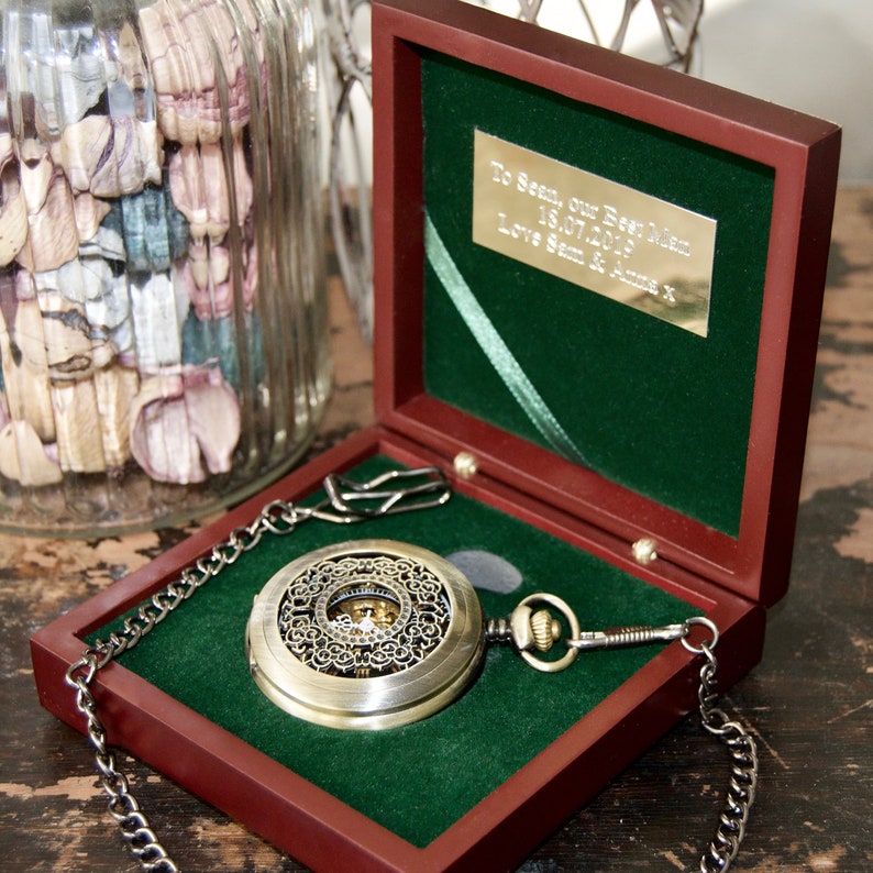 May include: A gold pocket watch with an intricate design and a chain, in a dark wood box with a green velvet lining. The box has a silver plaque with the inscription "To Sean, Our Dear Adam 18.07.2019 Love Sam & Anna X".