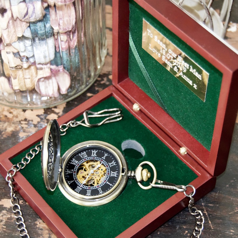 May include: A gold pocket watch with a black face and Roman numeral markings. The watch is open and the intricate inner workings are visible. The watch is in a red velvet box with a green velvet lining. The box has a gold plaque with the inscription "To Sean, our Best Man 18.07.2019 Love Sam & Anna x".