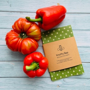 May include: Green and white polka dot beeswax food wrap with a brown label that reads "Knotty Bee Beeswax Food Wraps" and the instructions "Hand wash in cold water with alcohol-free soap" and "Avoid direct contact with meat". Three red bell peppers and a red tomato are on a light blue wooden surface.