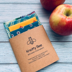 May include: A set of three beeswax food wraps in a brown paper envelope. The wraps are in different patterns: blue and yellow, yellow and blue, and blue and red. The envelope has the text "Knotty Bee Beeswax Food Wraps" and instructions for hand washing and avoiding direct contact with meat.