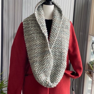 May include: A chunky, hand-knitted infinity scarf in grey, green, and brown hues, shown on a mannequin. The scarf features a textured knit and is styled over a red coat. A warm and stylish accessory for the colder months.