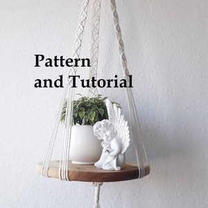 May include: A white macrame plant hanger with a wooden shelf, featuring a small white angel figurine and a potted plant. The hanger is suspended from the ceiling and has a tassel at the bottom. The text "Pattern and Tutorial" is visible in the image.