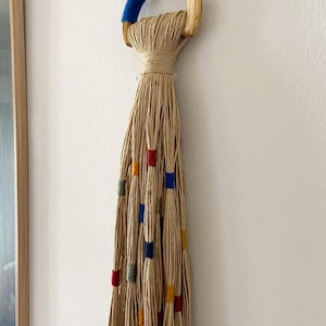 May include: A beige macrame plant hanger with a wooden ring top and colorful accents. The top ring is wrapped with blue fabric. The hanging strands are adorned with red, green, yellow, and blue square beads.