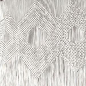 May include: A white macrame wall hanging with a geometric diamond pattern. The hanging has a fringe border.