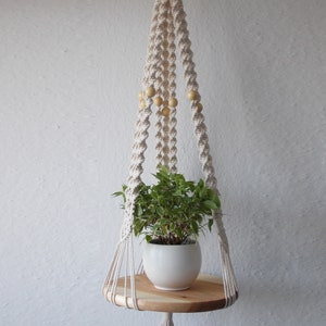 Può includere: Un portapiante in macramè con un ripiano rotondo in legno e un vaso in ceramica bianco con una pianta verde. La corda beige è decorata con perline di legno. Il portapiante è sospeso contro un muro bianco.