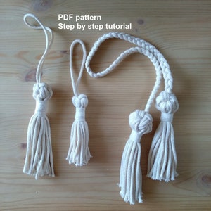 May include: Three white cotton tassels with a braided rope loop. The tassels are different sizes and are arranged in a row on a wooden surface. The text "PDF pattern Step by step tutorial" is written above the tassels.