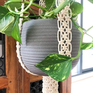 Pu&ograve; includere: Un vaso in ceramica grigia con una pianta verde appesa a un portavasi in macram&egrave; bianco.