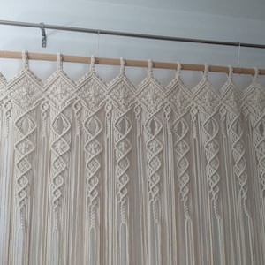 May include: Cream-colored macrame curtain with a geometric diamond pattern across the top and vertical strands. The curtain hangs from a wooden rod and metal brackets, adding a bohemian touch to home decor.