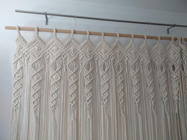 May include: Cream-colored macrame curtain with a geometric diamond pattern across the top and vertical strands. The curtain hangs from a wooden rod and metal brackets, adding a bohemian touch to home decor.