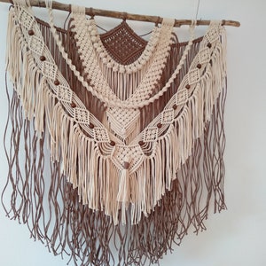 May include: A beige and brown macrame wall hanging with wooden beads and fringe. The hanging is made of cotton cord and is attached to a natural branch.