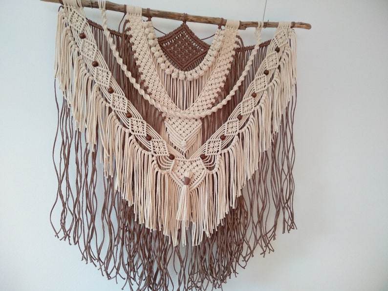 May include: A beige and brown macrame wall hanging with wooden beads and fringe. The hanging is made of cotton cord and is attached to a natural branch.