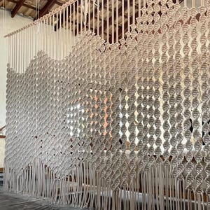 May include: A large macrame wall hanging made of natural beige cotton cord. The wall hanging features a repeating pattern of knots and is finished with long fringe.
