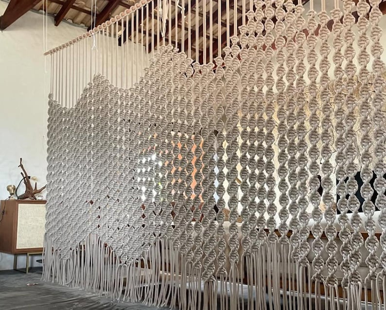 May include: A large macrame wall hanging made of natural beige cotton cord. The wall hanging features a repeating pattern of knots and is finished with long fringe.