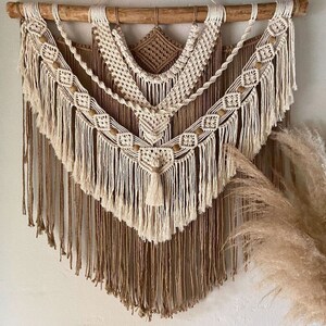 May include: A beige and white macrame wall hanging with wooden dowel and fringe. The wall hanging features a geometric pattern with wooden beads.