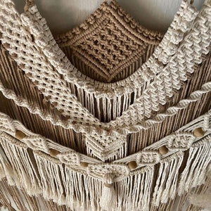 May include: A macrame wall hanging with a brown and white color scheme. The hanging features a geometric pattern with wooden beads and tassels.