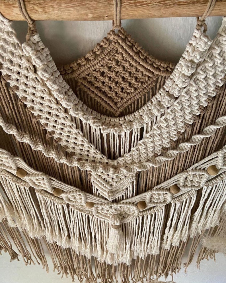 May include: A macrame wall hanging with a brown and white color scheme. The hanging features a geometric pattern with wooden beads and tassels.