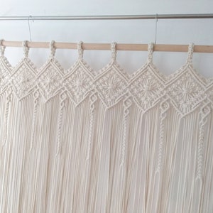 May include: A white macrame wall hanging with a geometric pattern and fringe. The hanging is made of cotton cord and is attached to a wooden dowel rod.