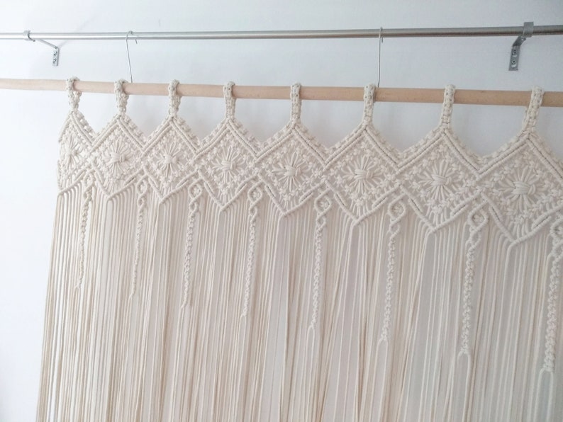 May include: A white macrame wall hanging with a geometric pattern and fringe. The hanging is made of cotton cord and is attached to a wooden dowel rod.