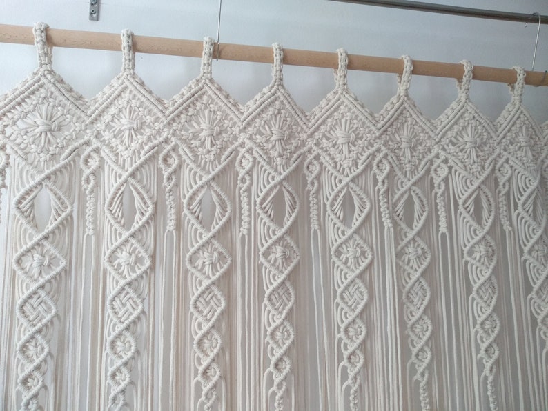 May include: Cream-colored macrame curtain with intricate geometric patterns. The curtain is suspended from a wooden rod and features diamond and floral designs. The macrame is made of thick, woven cotton cords, creating a textured, bohemian aesthetic.