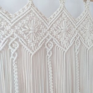 May include: A white macrame wall hanging with a geometric pattern. The hanging is made of cotton cord and is attached to a wooden dowel.
