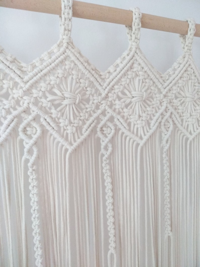 May include: A white macrame wall hanging with a geometric pattern. The hanging is made of cotton cord and is attached to a wooden dowel.