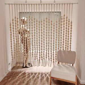 May include: A white macrame room divider with a woven rope design hangs from a white track. The divider is made of multiple strands of rope that are knotted together to create a pattern of circles. The divider is in front of a window with a white frame. A white chair with wooden legs sits in front of the divider.