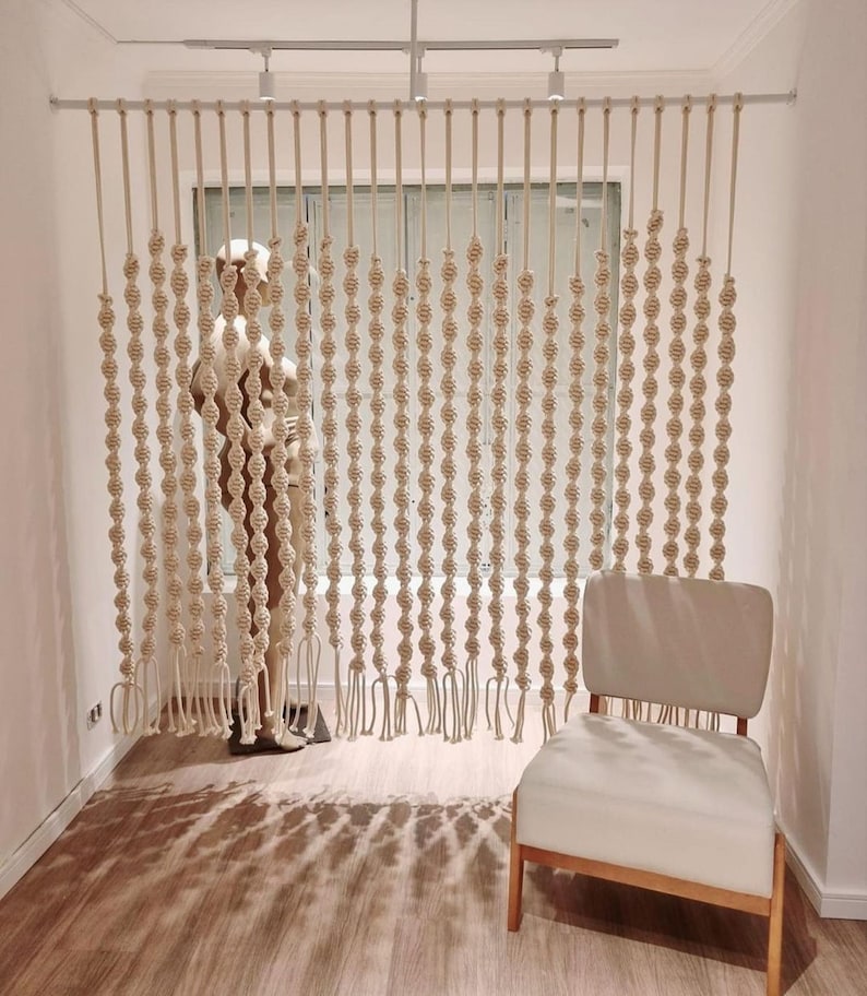 May include: A white macrame room divider with a woven rope design hangs from a white track. The divider is made of multiple strands of rope that are knotted together to create a pattern of circles. The divider is in front of a window with a white frame. A white chair with wooden legs sits in front of the divider.