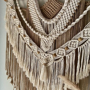 May include: A beige and brown macrame wall hanging with wooden beads and fringe. The hanging is made of natural fibers and has a bohemian style.