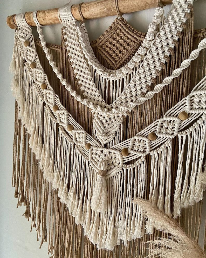 May include: A beige and brown macrame wall hanging with wooden beads and fringe. The hanging is made of natural fibers and has a bohemian style.