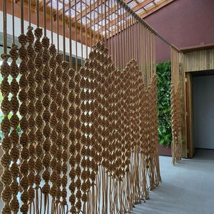 May include: A large macrame wall hanging made of natural brown rope. The wall hanging is made of a series of knots and is designed to resemble a curtain. The wall hanging is hanging in front of a wooden wall.